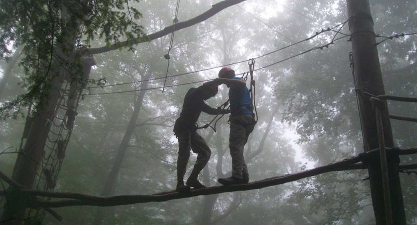 Two people wearing safety gear are secured by ropes as they navigate an obstacle of a high ropes course. The air around them appears foggy. 